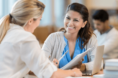two smiling women talking while one holds a pen and paper