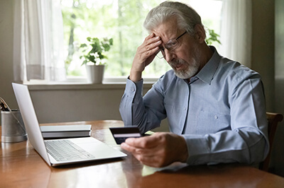 A grey haired man looking at his bank card in front of his laptop