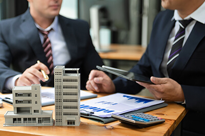 Men in suits looking at a calculator and charts. A model building is on the desk