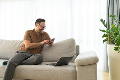man sitting on the couch looking at his smartphone with his laptop open
