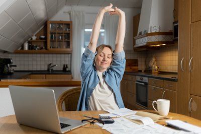 A woman sits at a table in the kitchen, happy from successfully completed financial work. There are papers, a laptop and a calculator on the table. success in financial management